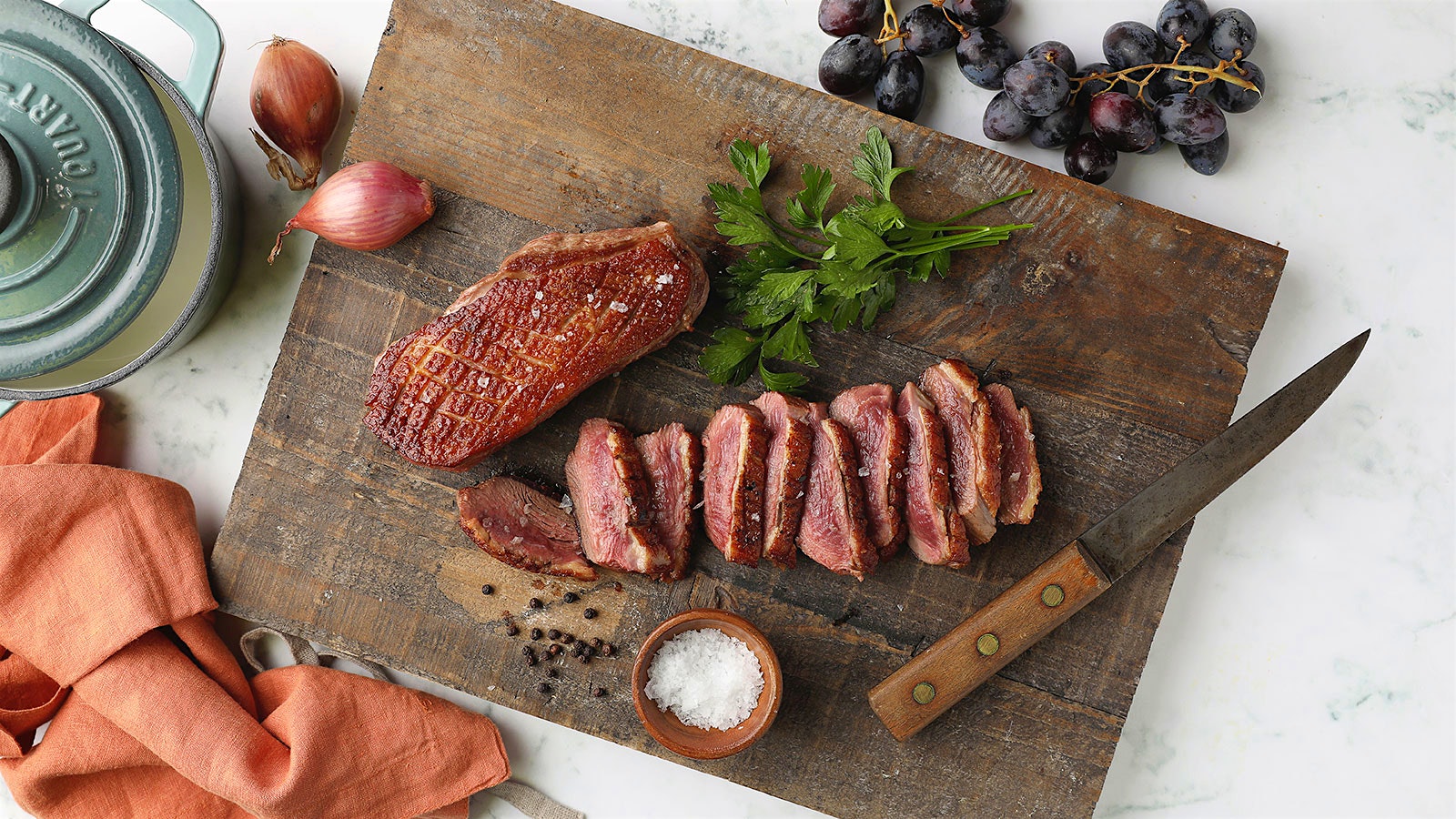 Sliced duck breast accompanied by garlic, parsley, a ramekin of salt, peppercorns and a bunch of red grapes